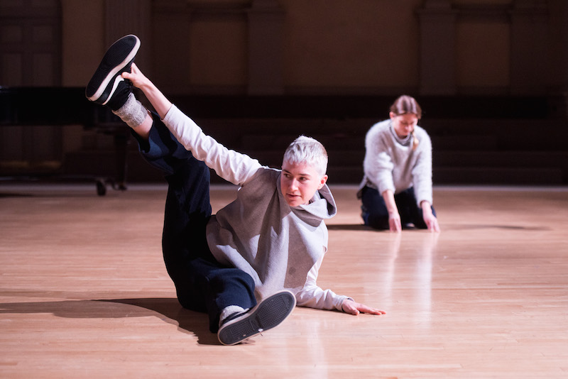 Two women sit on the floor in grey sweatshirts, blue pants and sneakers. The person in the foreground tips her hip to one side and holds on to her foot in the air
